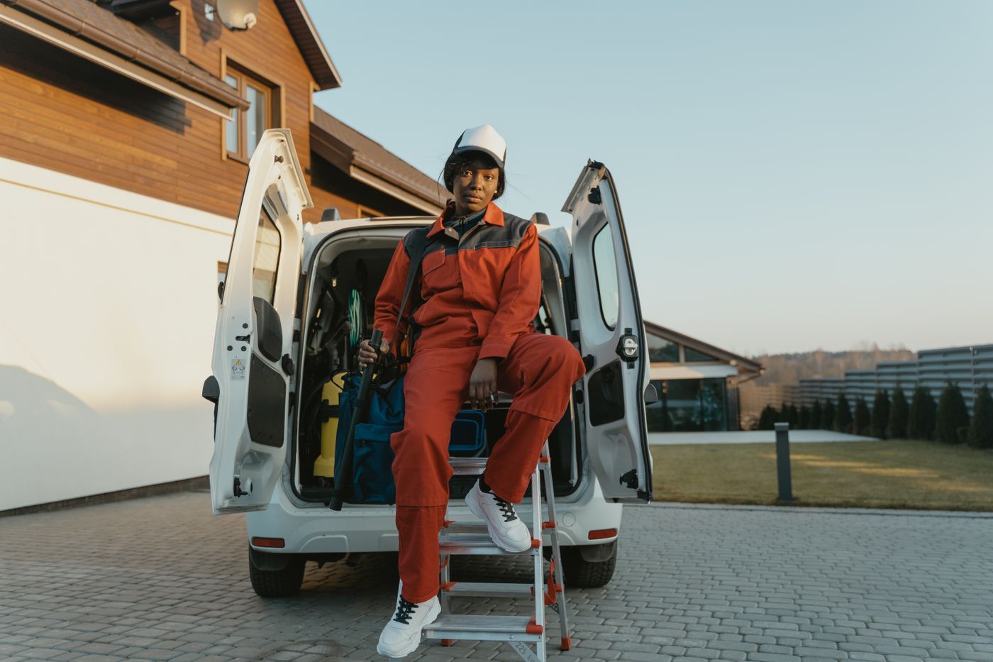 A worker in coveralls and a cap sitting beside an open service van on a paved driveway
