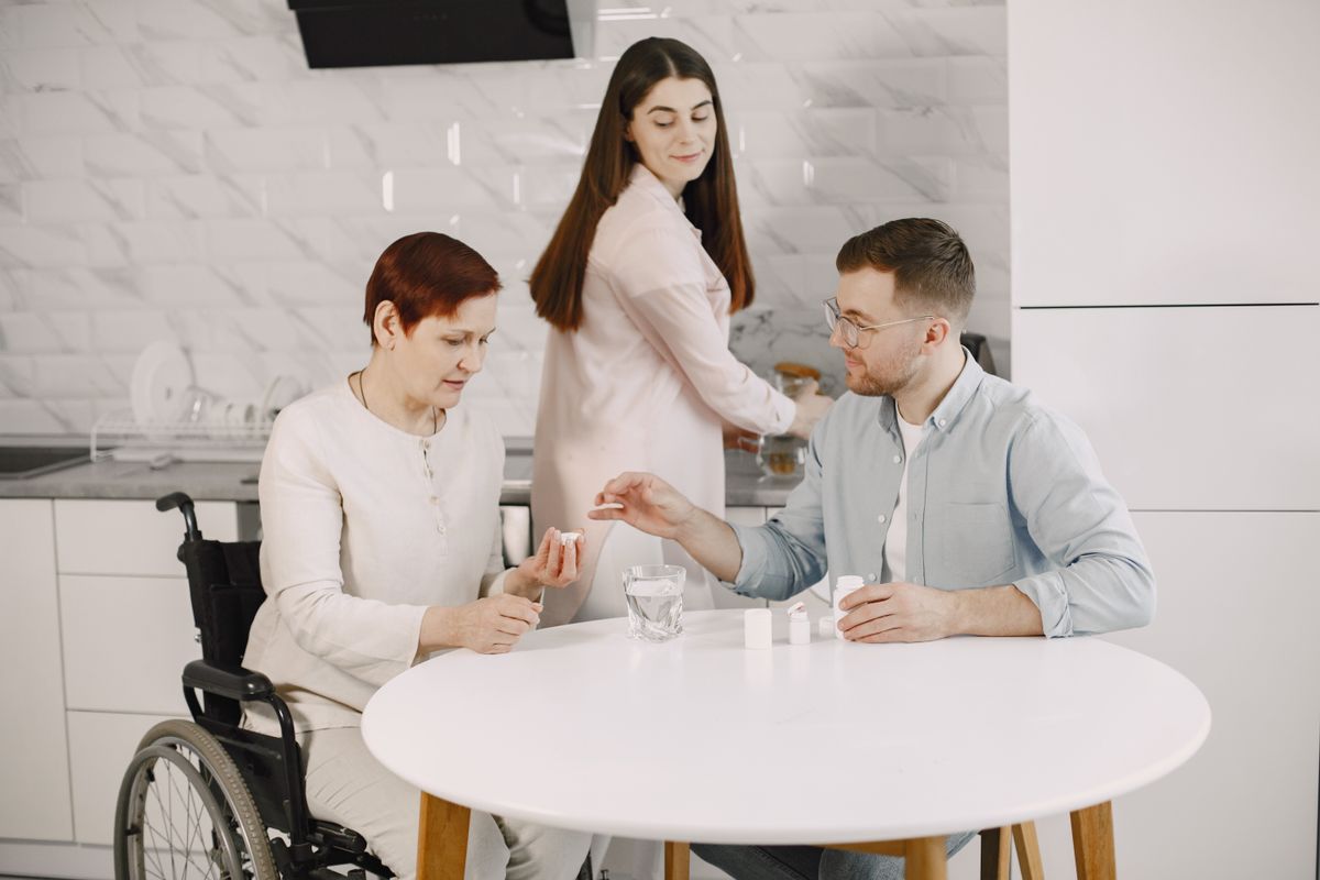 A woman in a wheelchair at a white kitchen table with two younger adults and small medicine bottles, one handing her a pill