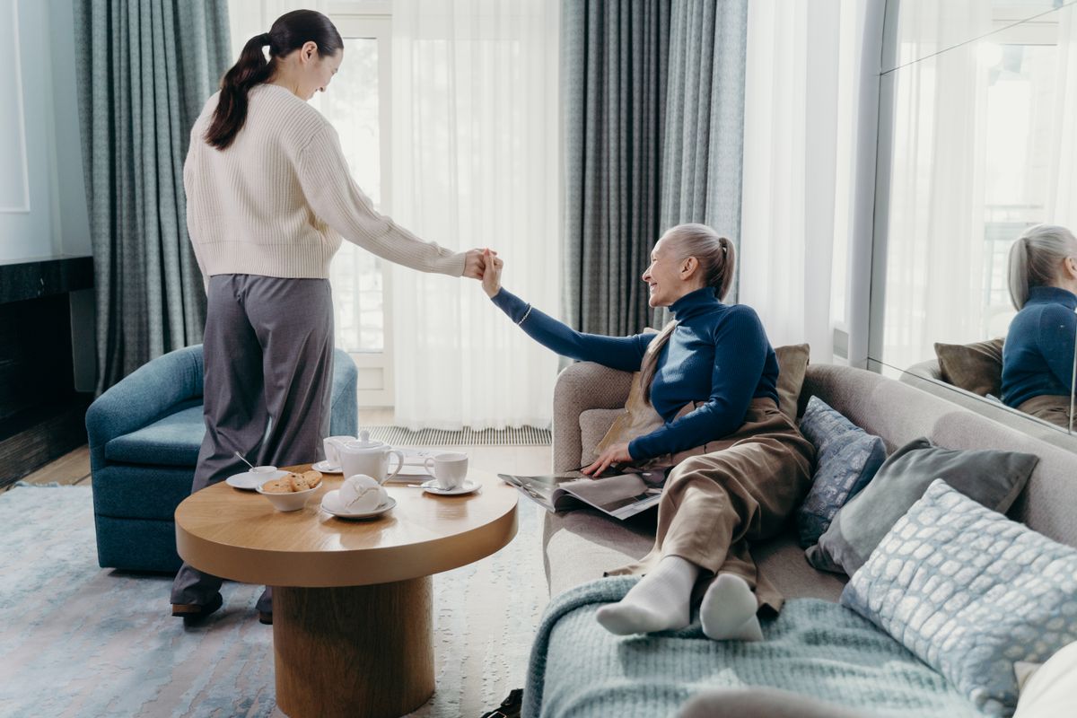 An older woman on a sofa smiling and holding hands with a younger woman in a bright living room, with tea on the table