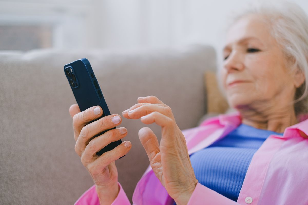An older woman with white hair sitting on a sofa and looking at a smartphone in her hands