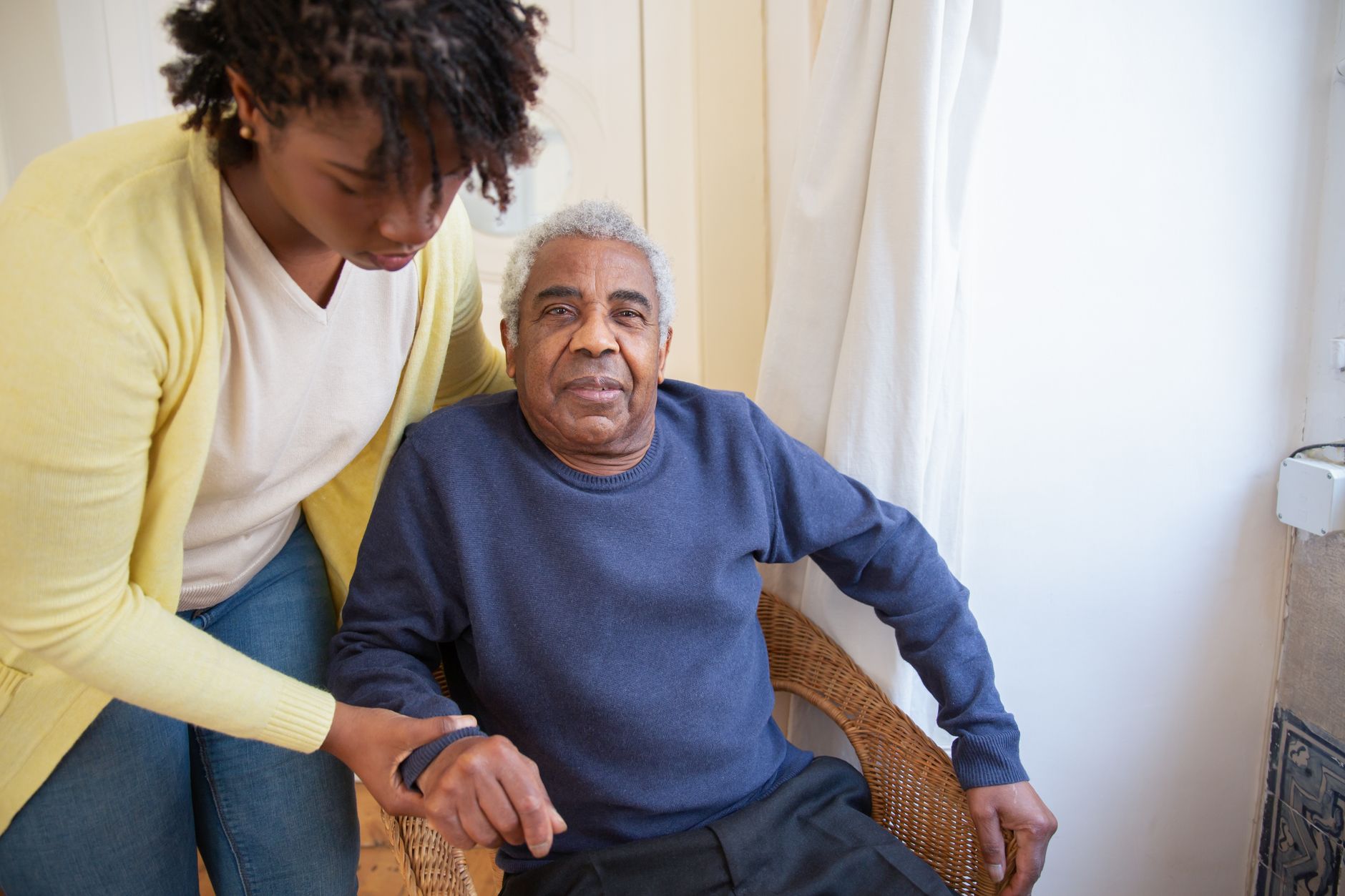 A caregiver helping an older man stand up in a bright room at home