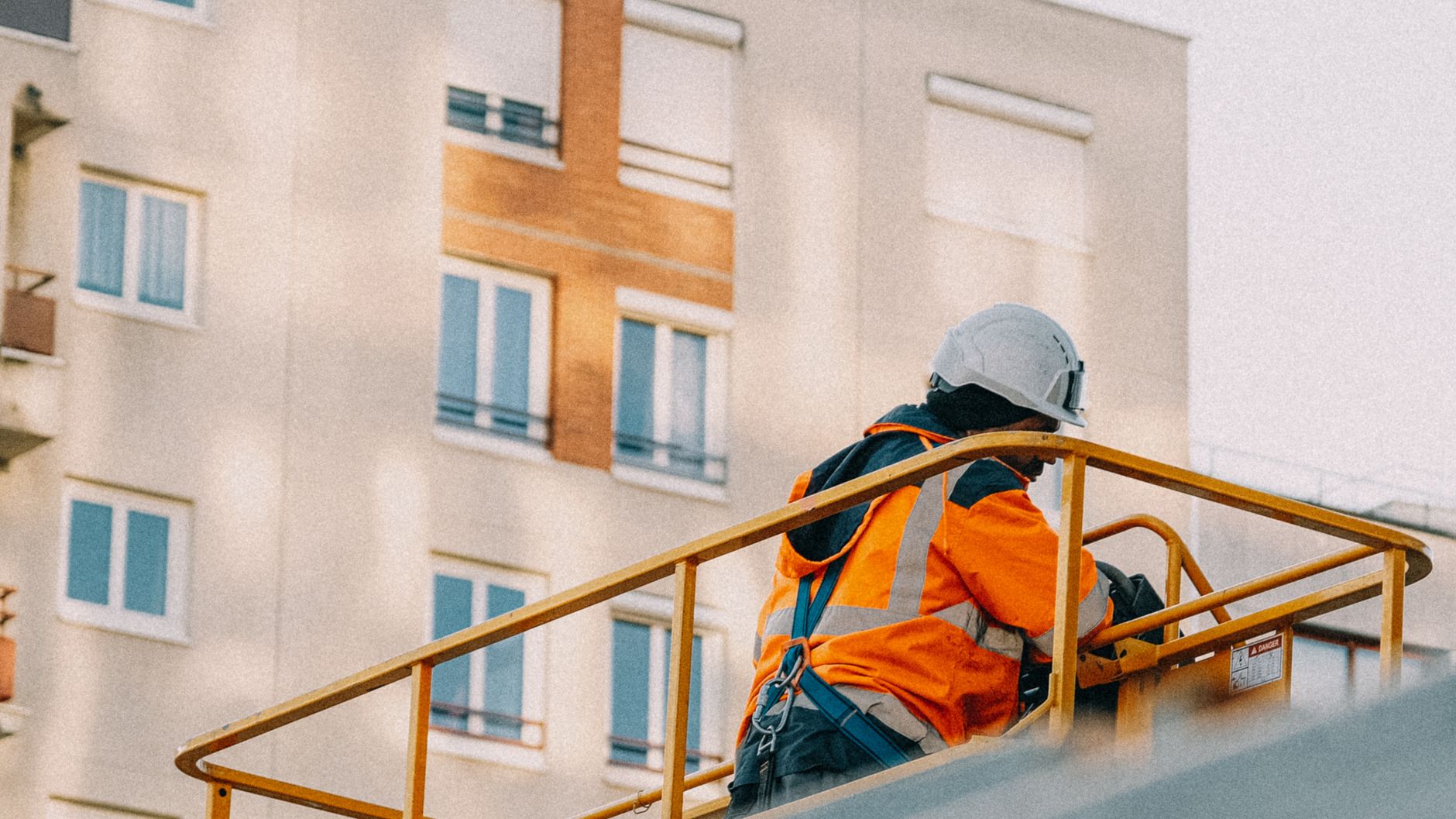 A worker in safety gear on a lift beside an apartment building exterior